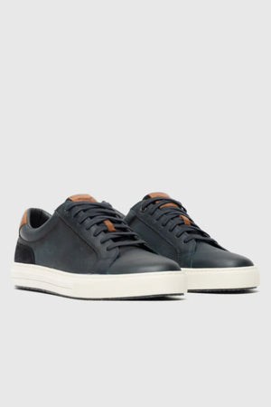 A pair of Rodd & Gunn Transit Sneakers in black with white soles, black laces, and tan leather accents on the tongue and heel are displayed on a light gray background.