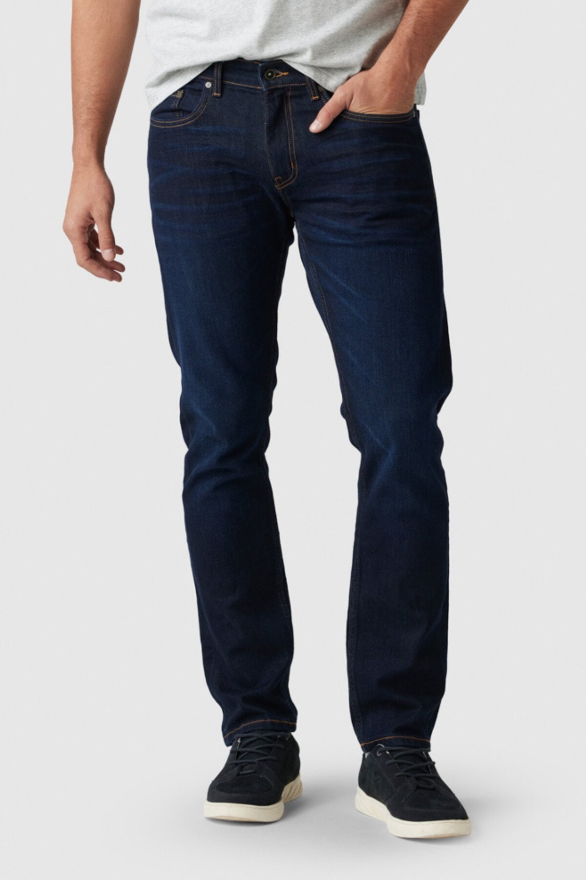 A man in a light gray t-shirt, Rodd & Gunn Fanshawe Straight Jeans, and black sneakers with white soles stands with one hand in his pocket against a plain light background.