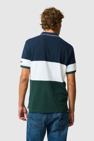 A person with curly hair wears a Rodd & Gunn Homebay Polo in a color-blocked navy, white, and green sports-fit style with short sleeves, paired with blue jeans, standing back to the camera against a plain light background.