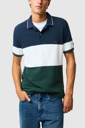 A person stands against a light gray background wearing the Rodd & Gunn Homebay Polo, made from Australian Good Earth Cotton, with navy, white, and green color-blocking, an embroidered chest logo, red-trimmed collar, and blue jeans.