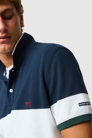 A man wears the Rodd & Gunn Homebay Polo in navy and white sports fit, featuring green trim, a red dog logo, and a "RODD & GUNN" sleeve patch. Made from Australian Good Earth Cotton. The background is plain light gray.