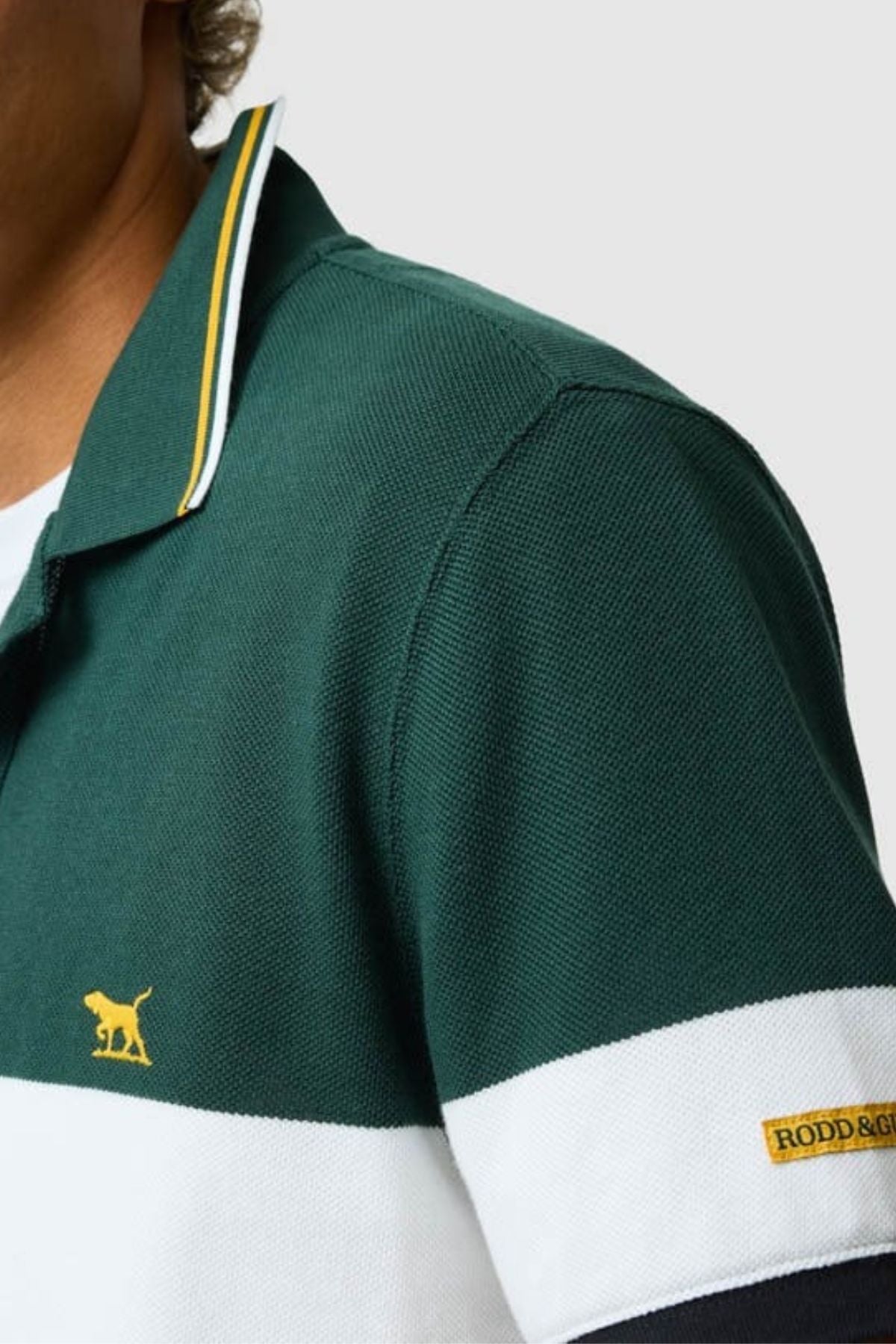 Close-up of a person wearing the green and white Rodd & Gunn Homebay Polo with a yellow animal logo and "RODD & GUNN" tag, crafted from sustainable Australian Good Earth Cotton. Set against a plain light gray background.