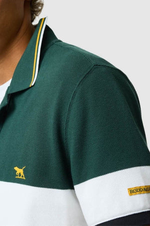 Close-up of a person wearing the green and white Rodd & Gunn Homebay Polo with a yellow animal logo and "RODD & GUNN" tag, crafted from sustainable Australian Good Earth Cotton. Set against a plain light gray background.