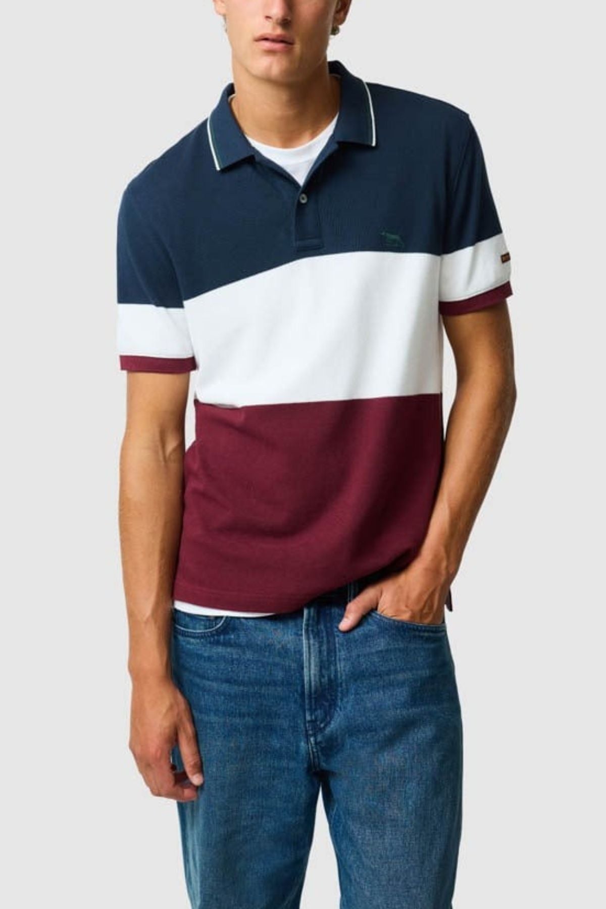 A man stands with one hand in his pocket against a plain light background, wearing the Rodd & Gunn Homebay Polo—a breathable pique knit short-sleeve shirt with navy, white, and burgundy color blocks—paired with blue jeans.