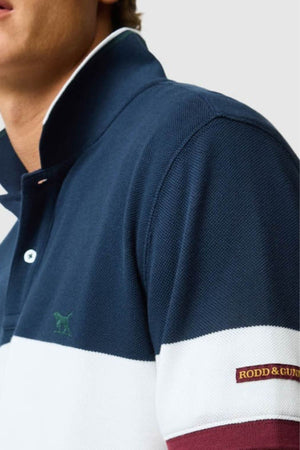 A person wears the Rodd & Gunn Homebay Polo—navy blue and white, collared, with a small green dog logo on the chest and "RODD & GUNN" patch on the sleeve—crafted from breathable pique knit. Only their lower face and upper torso are visible.