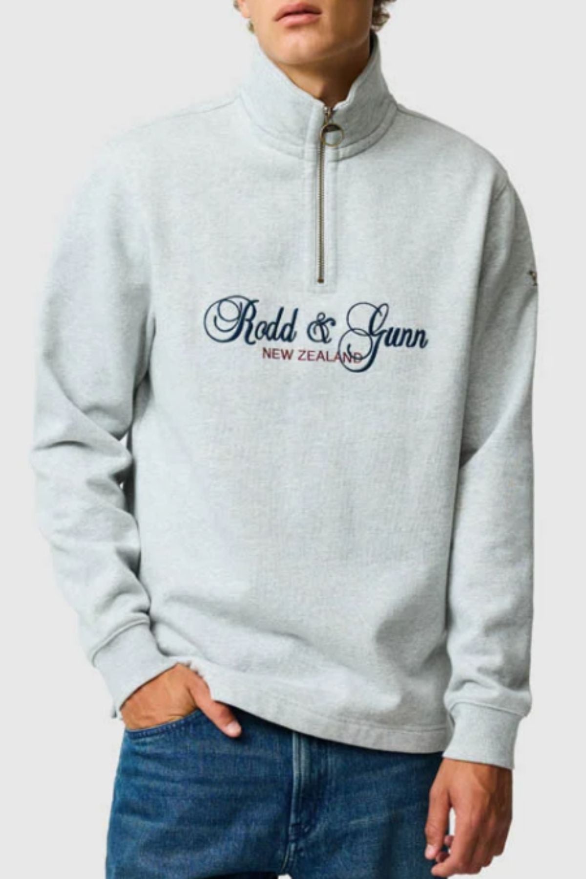 A person wearing the Rodd & Gunn Jubilee Sweat, a light grey quarter zip with "Rodd & Gunn New Zealand" in dark blue on the front, paired with blue jeans and one hand in their pocket.