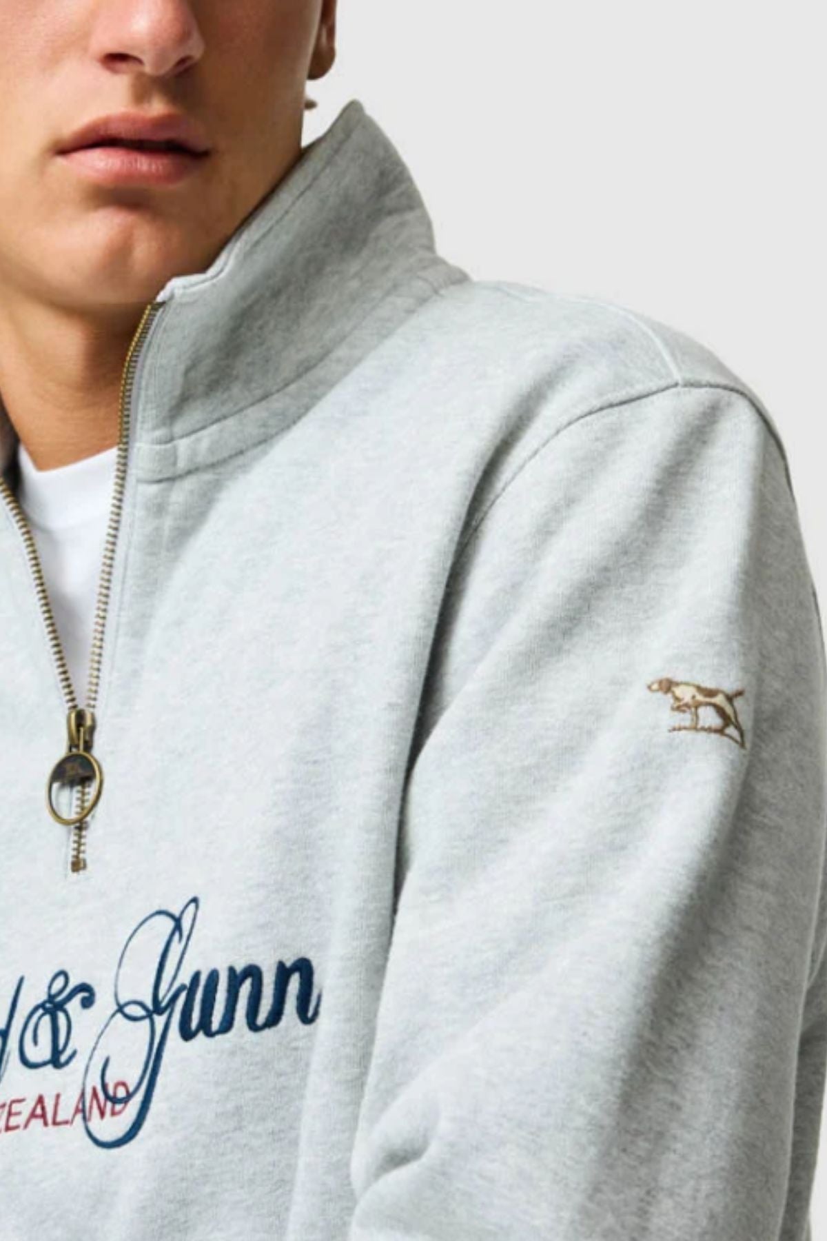 A close-up of a person wearing the Rodd & Gunn Jubilee Sweat in light gray, featuring a gold zipper, blue embroidered text, and a small embroidered dog on the sleeve—showing only their lower face and upper torso.