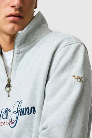 A close-up of a person wearing the Rodd & Gunn Jubilee Sweat in light gray, featuring a gold zipper, blue embroidered text, and a small embroidered dog on the sleeve—showing only their lower face and upper torso.