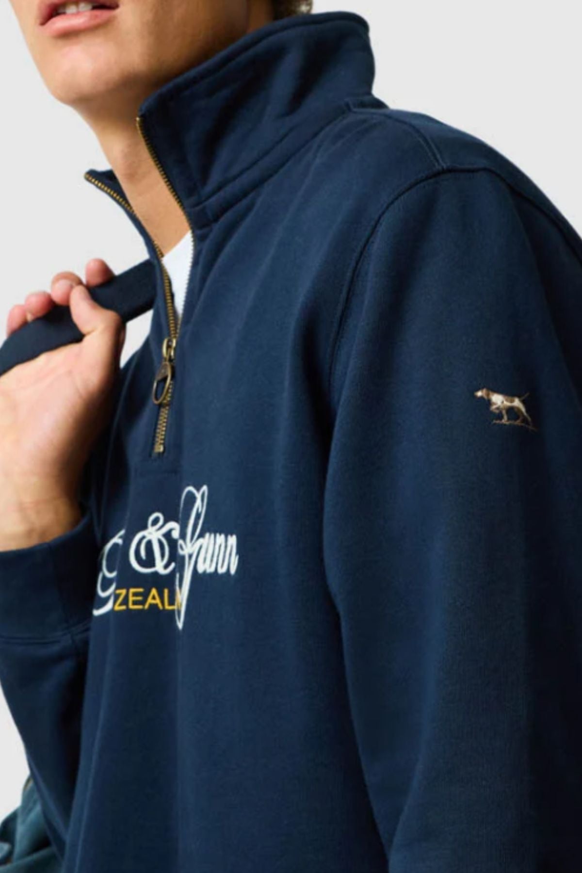 A person wearing the Rodd & Gunn Jubilee Sweat in navy, made with Australian Good Earth Cotton, featuring white and yellow embroidered text and a small dog logo on the sleeve, holds the collar strap with one hand.