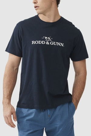 A man is wearing a dark blue Rodd & Gunn Logo T-Shirt, crafted from sustainably farmed cotton, featuring a small graphic of a dog. He pairs it with blue shorts and looks away from the camera, his right hand casually resting near his hip against a plain white background.