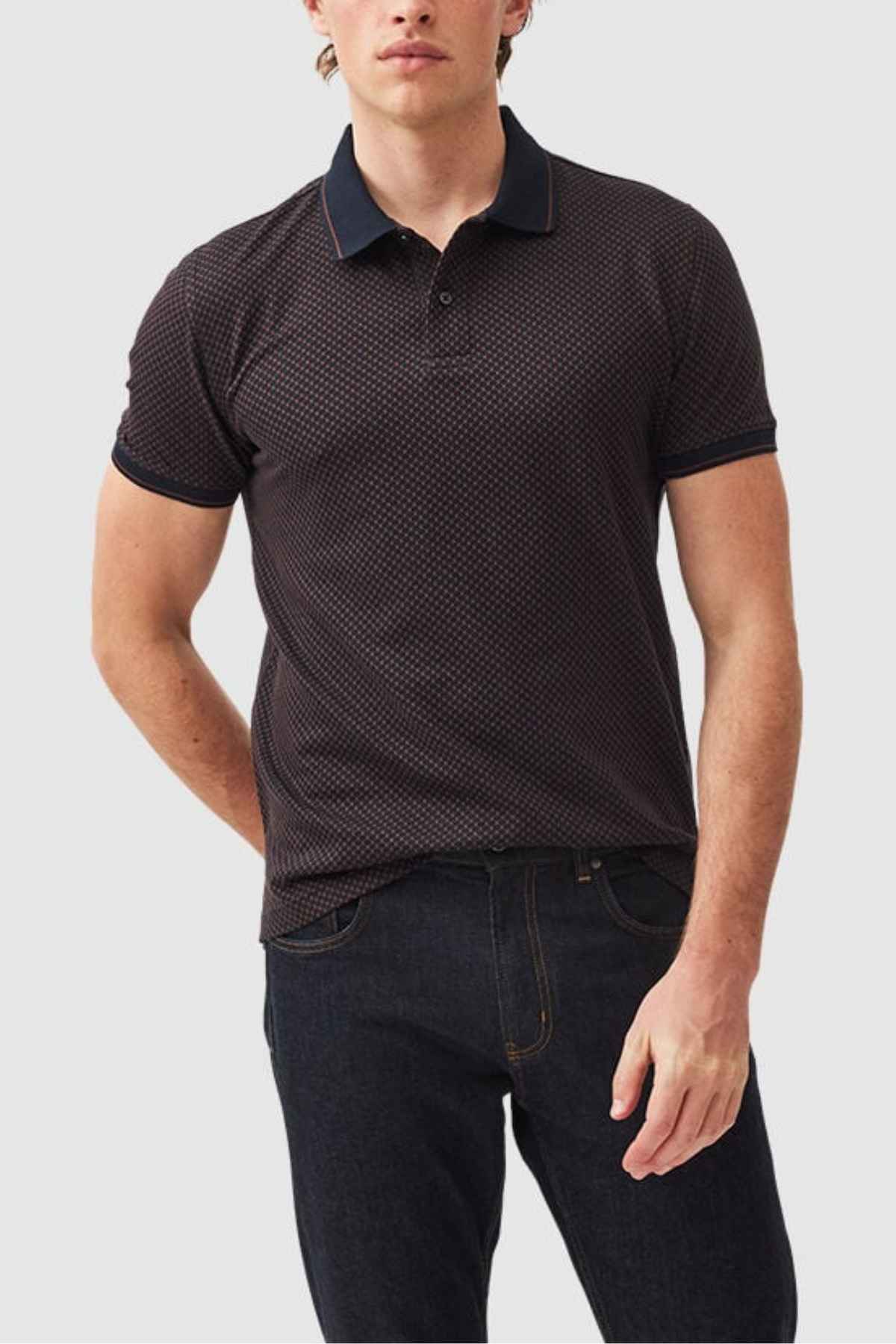 A man stands against a plain background, wearing the Rodd & Gunn Lynden Sports Fit Polo made from premium cotton yarn and dark jeans. His right hand slips casually into his pocket while his left arm rests elegantly at his side.