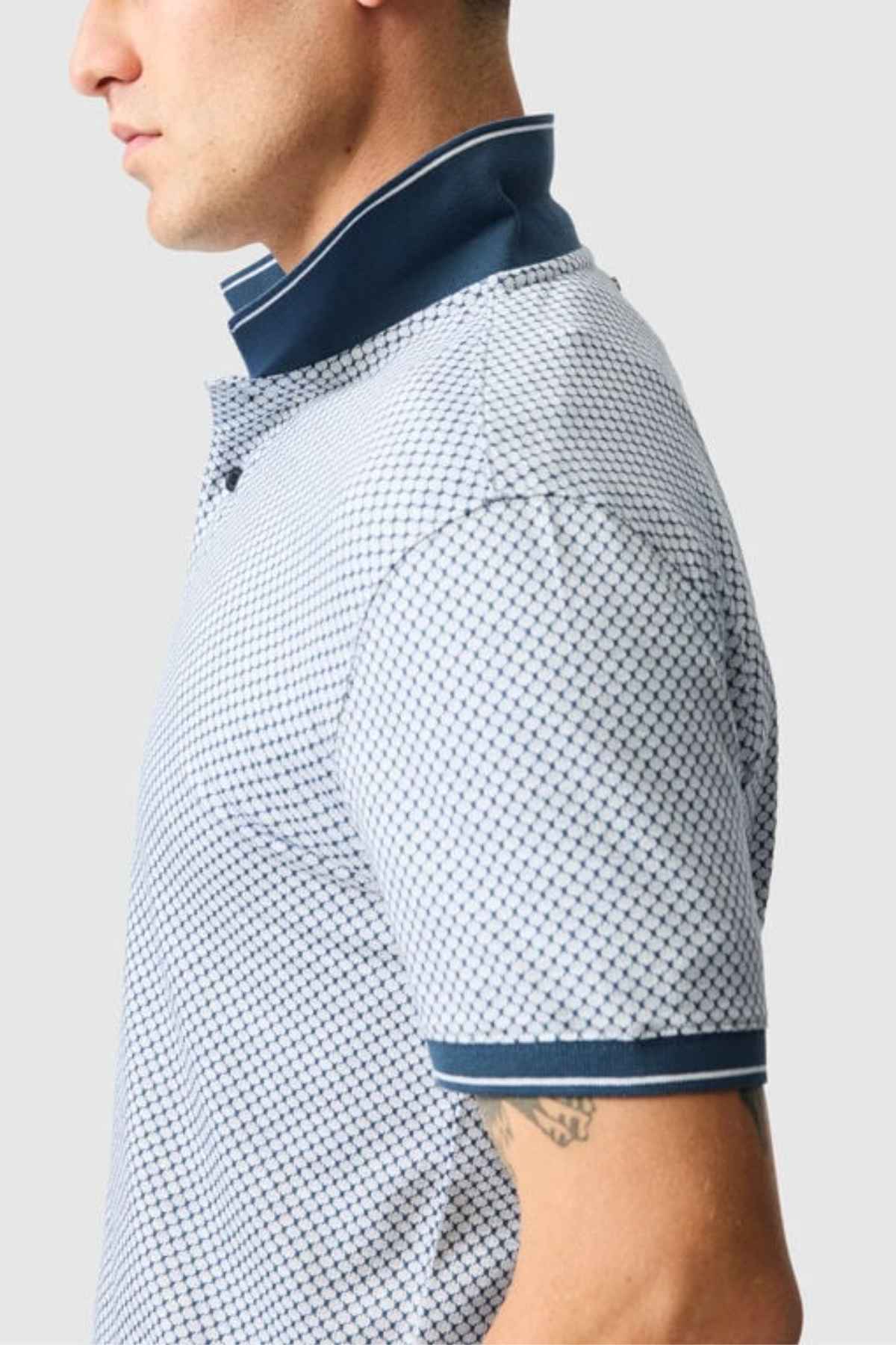 A person wearing the Rodd & Gunn Lynden Sports Fit Polo in light blue, accented with a dark blue collar and cuffs. Crafted from luxurious Turkish cotton, this shirt showcases small geometric shapes, enhancing their style. The collar is slightly turned up, revealing a tattoo on their left arm.