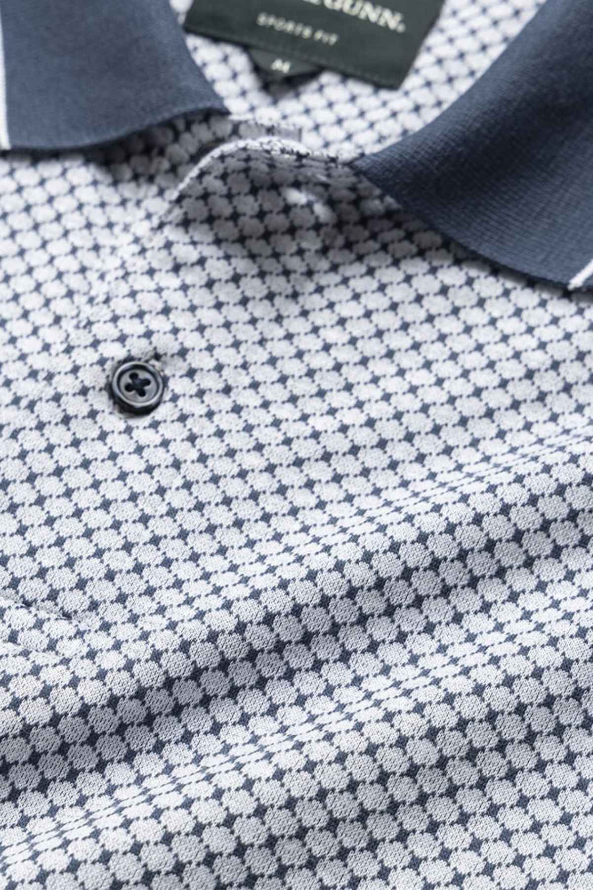 Close-up of a Rodd & Gunn Lynden Sports Fit Polo made from luxurious Turkish cotton with a small geometric pattern in navy and white. The shirt has a dark collar, a single visible button, and the tag is partially visible inside the collar.