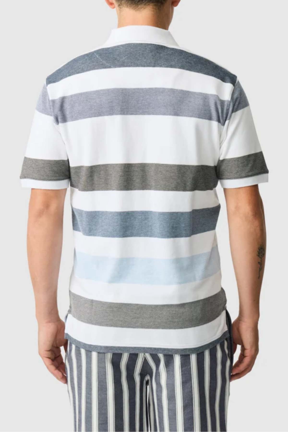 A person is wearing the Rodd & Gunn Milton Polo, featuring short sleeves and horizontal stripes in blue, grey, and white. They're also in grey and white vertically striped pants made of four-way stretch fabric, with the image captured from the back.