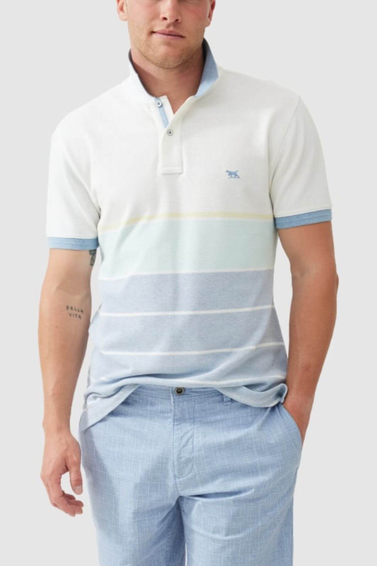 A man wears the Rodd & Gunn Mission Bay Sports Fit Polo, featuring bold blue and white horizontal stripes and four-way stretch fabric, paired with light blue pants. He stands against a plain gray background with his left hand in his pocket.