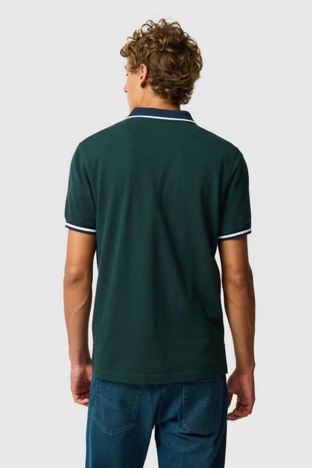 A man with curly hair, facing away from the camera, wears the Rodd & Gunn New Lynn Polo—sports fit with Australian Good Earth Cotton and white trim on the collar and sleeves—paired with blue jeans against a plain light background.
