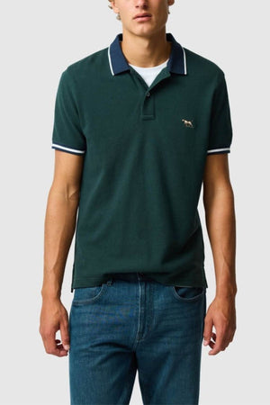 A person stands against a plain light gray background, wearing the Rodd & Gunn New Lynn Polo in dark green with white collar and sleeve trim, made from Australian Good Earth Cotton, paired with blue jeans.