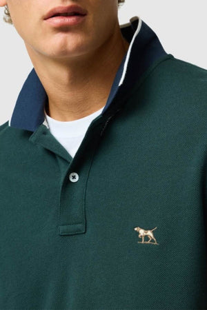 A person wearing a dark green Rodd & Gunn New Lynn Polo with a small embroidered dog logo on the chest and a white undershirt visible beneath the unbuttoned collar.