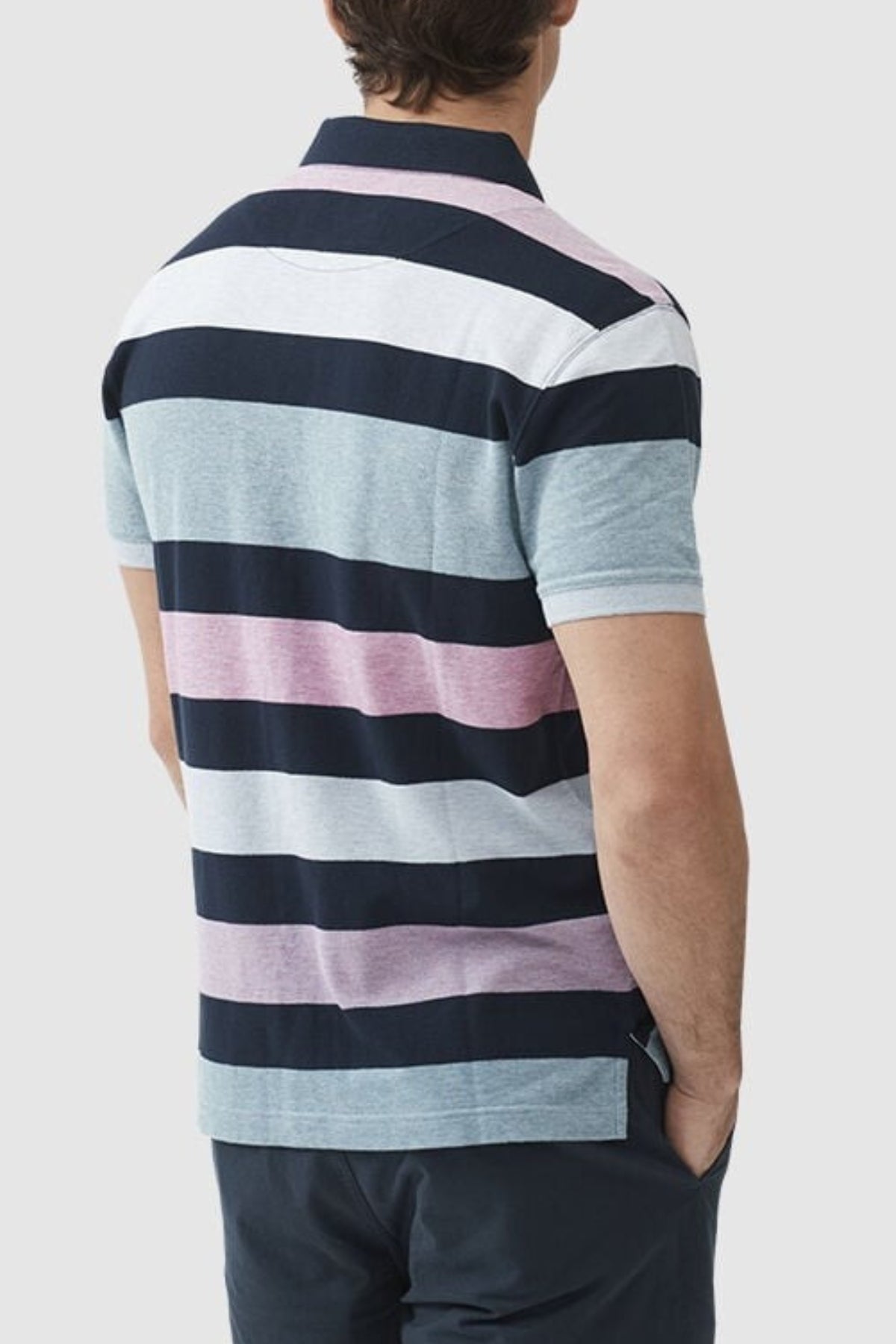 Someone stands with their back to the camera, wearing a Rodd & Gunn Solomons Point Sports Fit Polo with wide dark blue, pink, light gray, and white stripes. Made from 100% cotton with four-way stretch for comfort, they have one hand in their pocket against a plain light gray background.