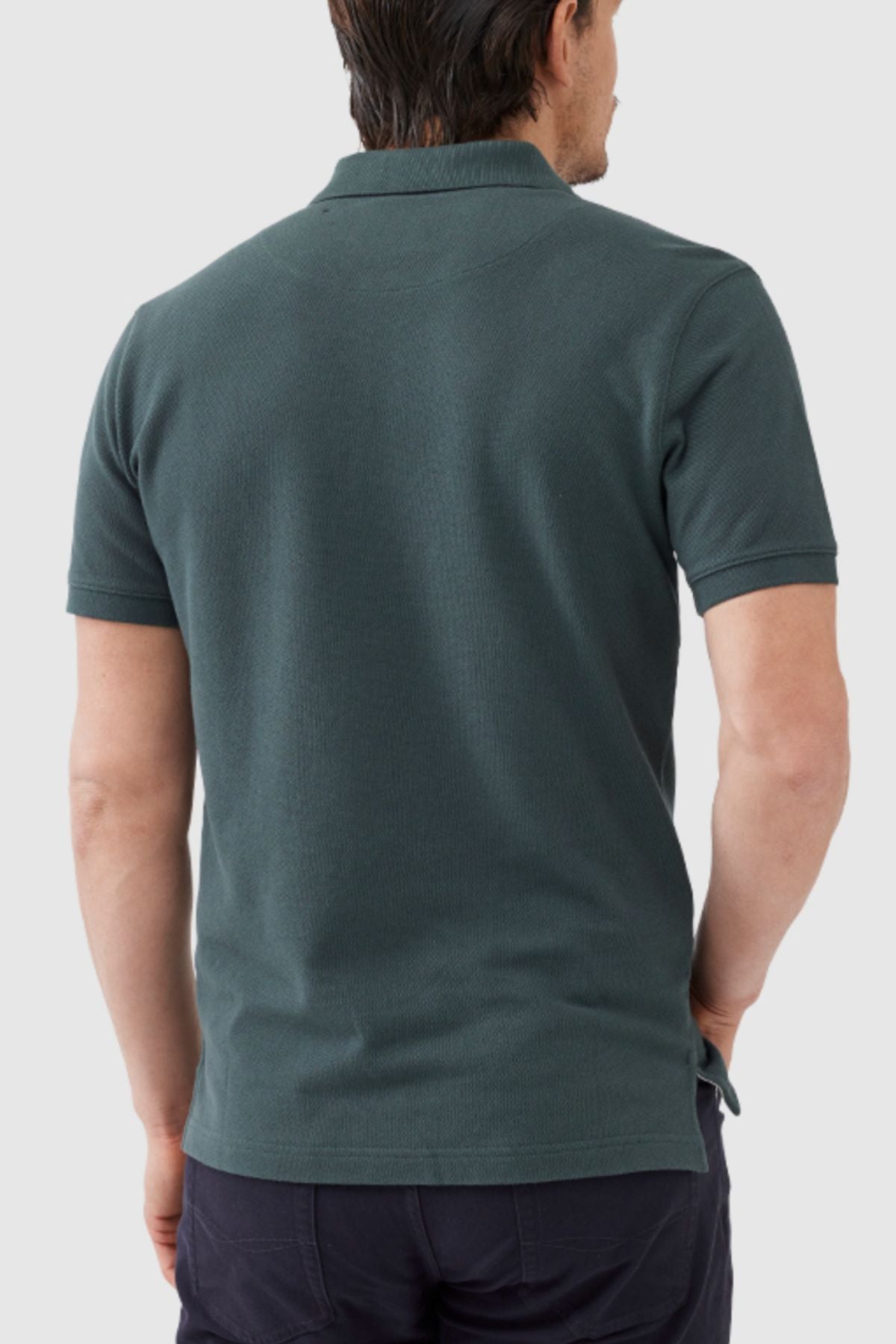 A man is shown from the back wearing a Rodd & Gunn The Gunn Polo in dark green, paired with dark pants. Made from premium Australian Cotton, the modern fit casual polo shirt accentuates his dark hair while his left hand rests in his pocket against a plain, light-colored background.