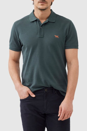 A man wearing the Rodd & Gunn The Gunn Polo in dark green, featuring a small red animal logo on the left chest pocket area. This modern-fit shirt, made from Australian cotton, is paired with dark trousers. He has one hand in his pocket against a light gray background. The man's face is partially visible, emphasizing the clothing.