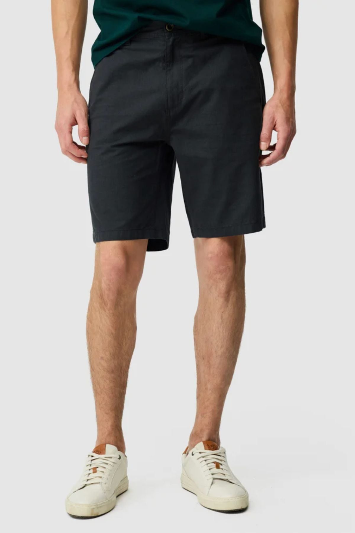 A person wears Rodd & Gunn The Gunn Short in classic fit cotton elastane, paired with a dark green shirt and white sneakers, standing against a plain light gray background. Only the body from the shoulders down is visible.