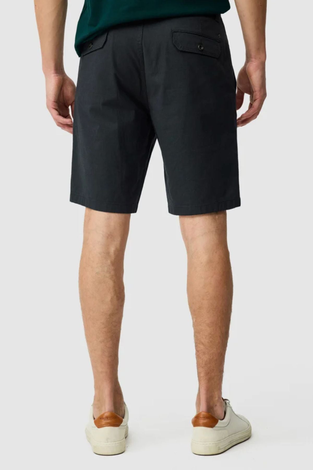 A man stands with his back to the camera, wearing Rodd & Gunn The Gunn Short in cotton-elastane with buttoned back pockets, paired with white sneakers featuring tan heel accents. The background is plain and light-colored.