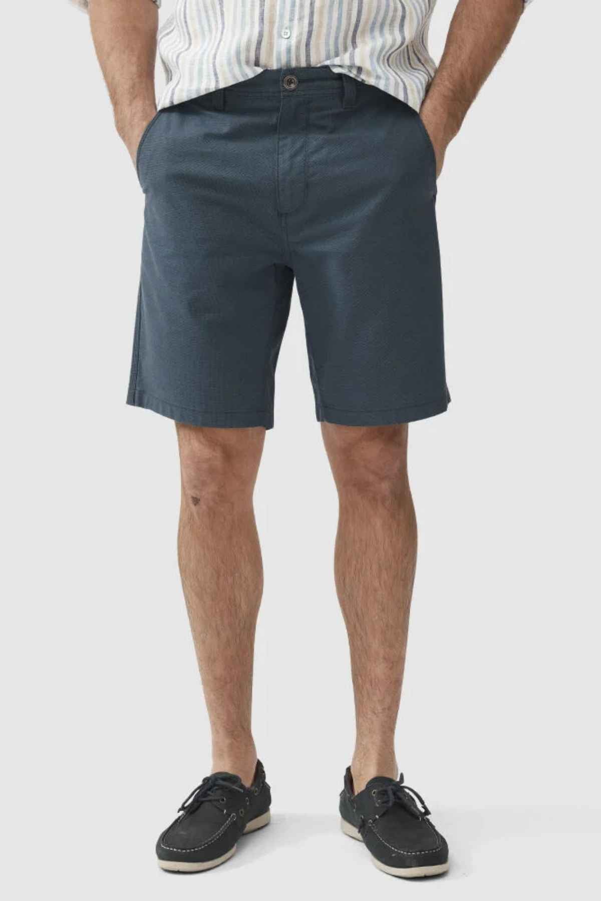 A person wearing a striped shirt, Rodd & Gunn The Gunn Short in an original fit made from high-quality teal cotton, and black loafers stands with hands in pockets. The image captures the lower half of the body against a plain background.