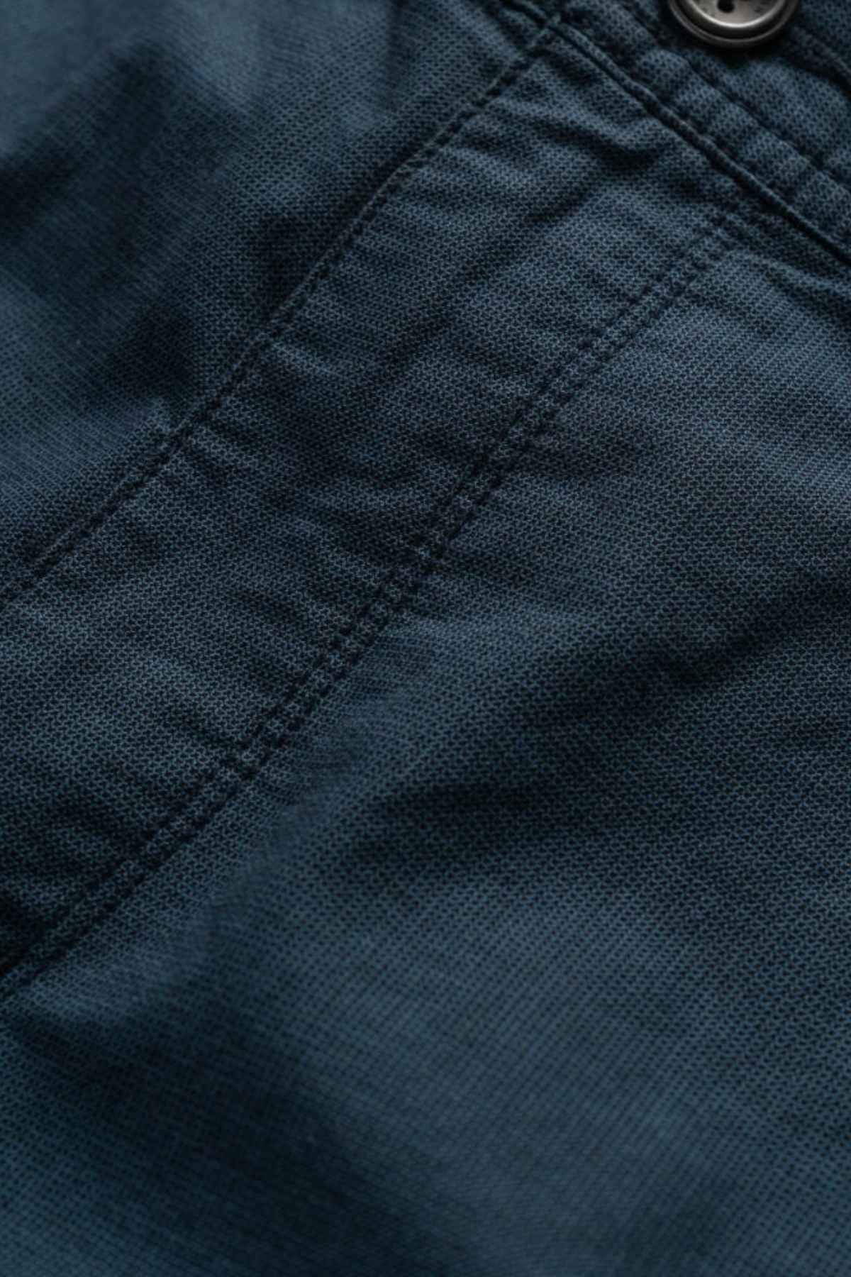 Close-up of the Rodd & Gunn The Gunn Short's dark fabric, showcasing a visible stitched seam with a button at the top right. The high-quality cotton texture appears coarse, hinting at its durable material. The seam runs vertically through the center, emphasizing its original fit design.