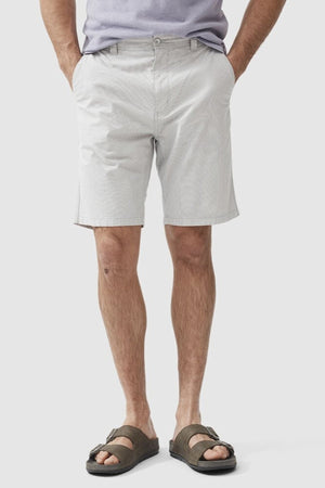 A man, with only his lower half visible against a plain white background, is wearing Rodd & Gunn The Gunn Short in light gray, made from a cotton elastane blend. He has his hands in the pockets of the classic fit shorts and is also donning greenish-brown sandals. Additionally, he appears to be wearing a light-colored shirt.