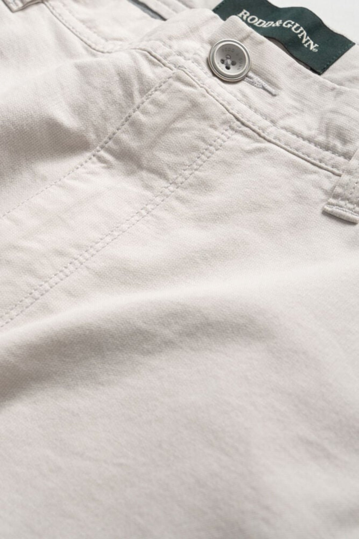 Close-up image of light beige Rodd & Gunn The Gunn Short pants with visible stitching, button, and belt loop, featuring a Rodd & Gunn label on the inner waistband. These classic fit shorts are crafted from a cotton elastane blend for added comfort and durability.