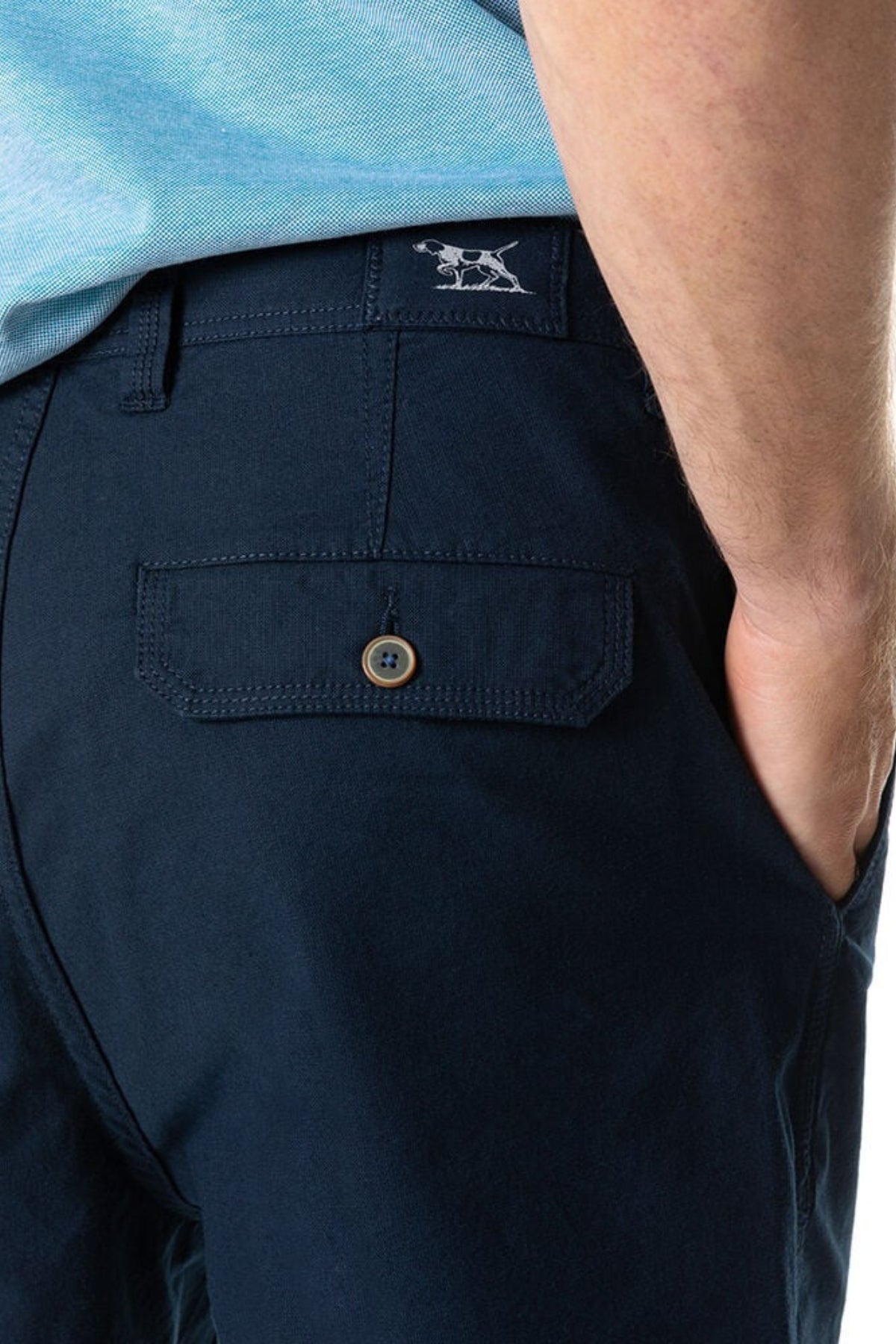 A close-up of a person wearing Rodd & Gunn The Gunn Short in dark blue, featuring a buttoned back pocket. The person's hand is partially visible, resting in the short's pocket. The top of the shorts displays a stitched Rodd & Gunn logo near the waistband, and they are paired with a light blue shirt.