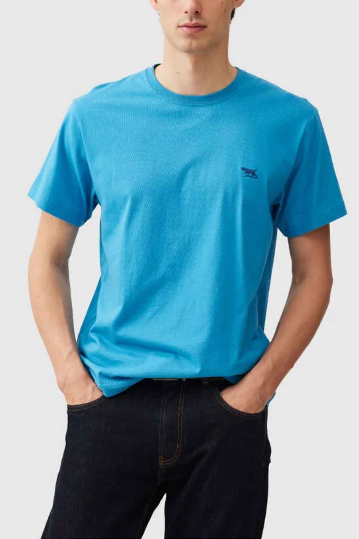 Someone stands with hands in pockets against a light gray background, wearing a plain blue Rodd & Gunn The Gunn T-Shirt made from Australian cotton and dark jeans.