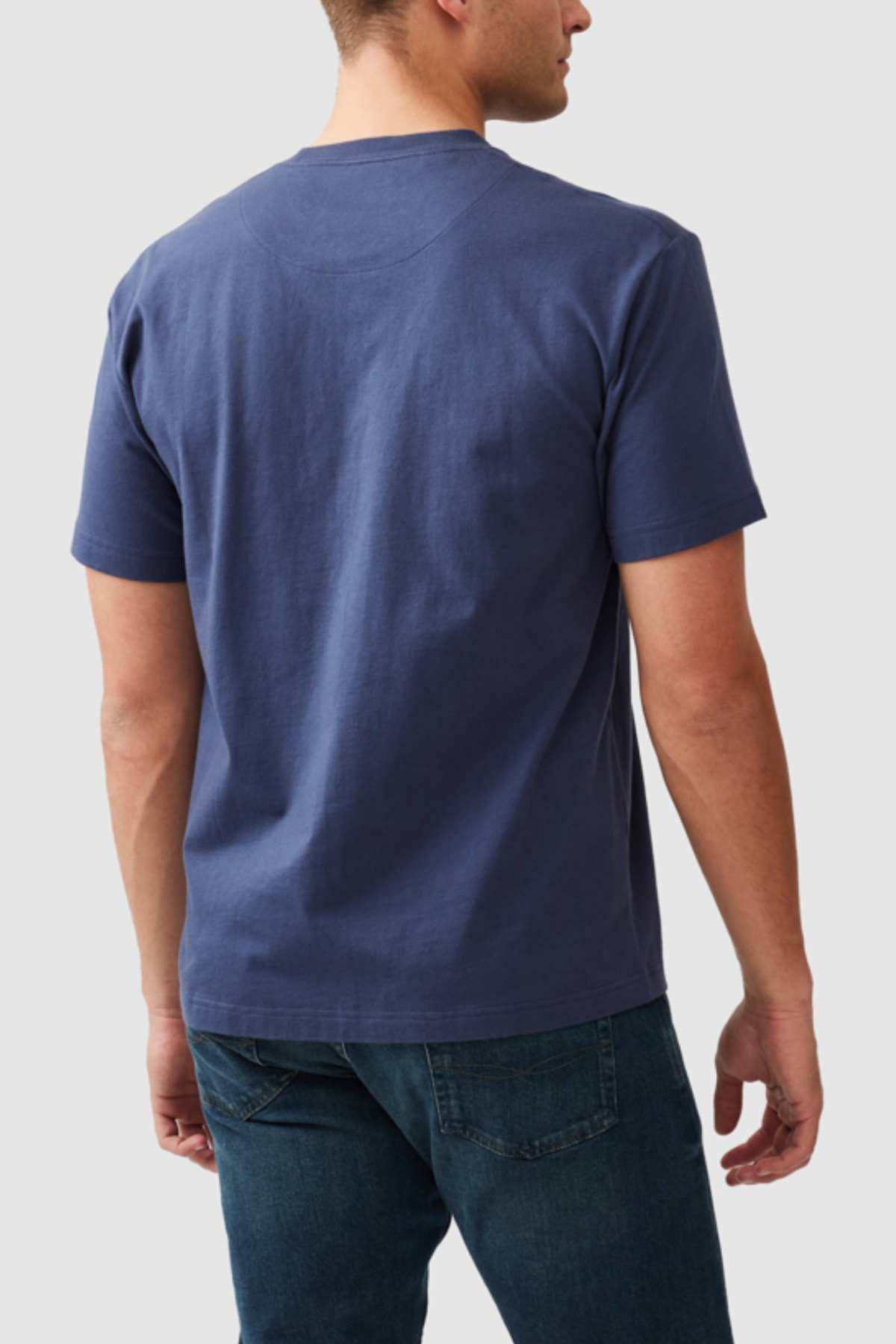 A person stands facing away from the camera, wearing a Rodd & Gunn The Peaks Logo Tee and blue jeans. The background is light gray. The person's left arm is slightly bent, and they have short hair.