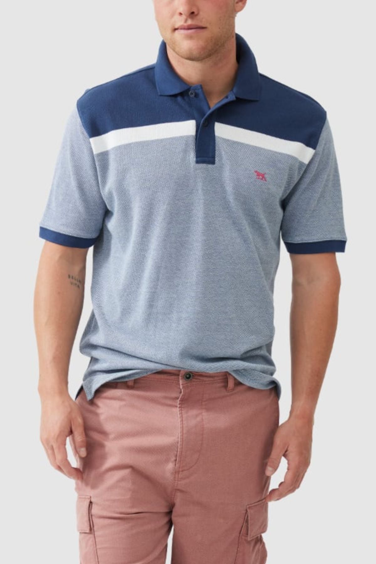 A man stands against a plain background, wearing a Rodd & Gunn Whitby Polo featuring a bold color block design with white and navy stripes, paired with dusty rose cargo shorts.