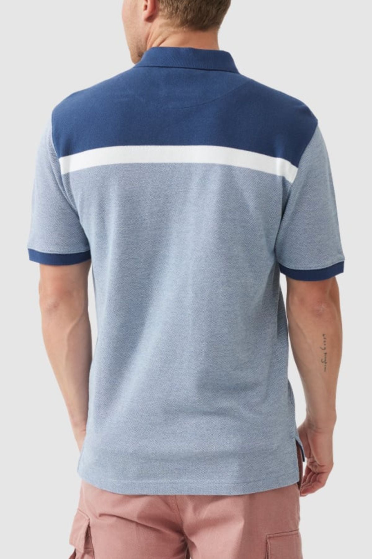 A person sporting the Rodd & Gunn Whitby Polo with a bold blue and gray color block design and a white horizontal stripe across the back stands facing away, wearing vibrant pink shorts.