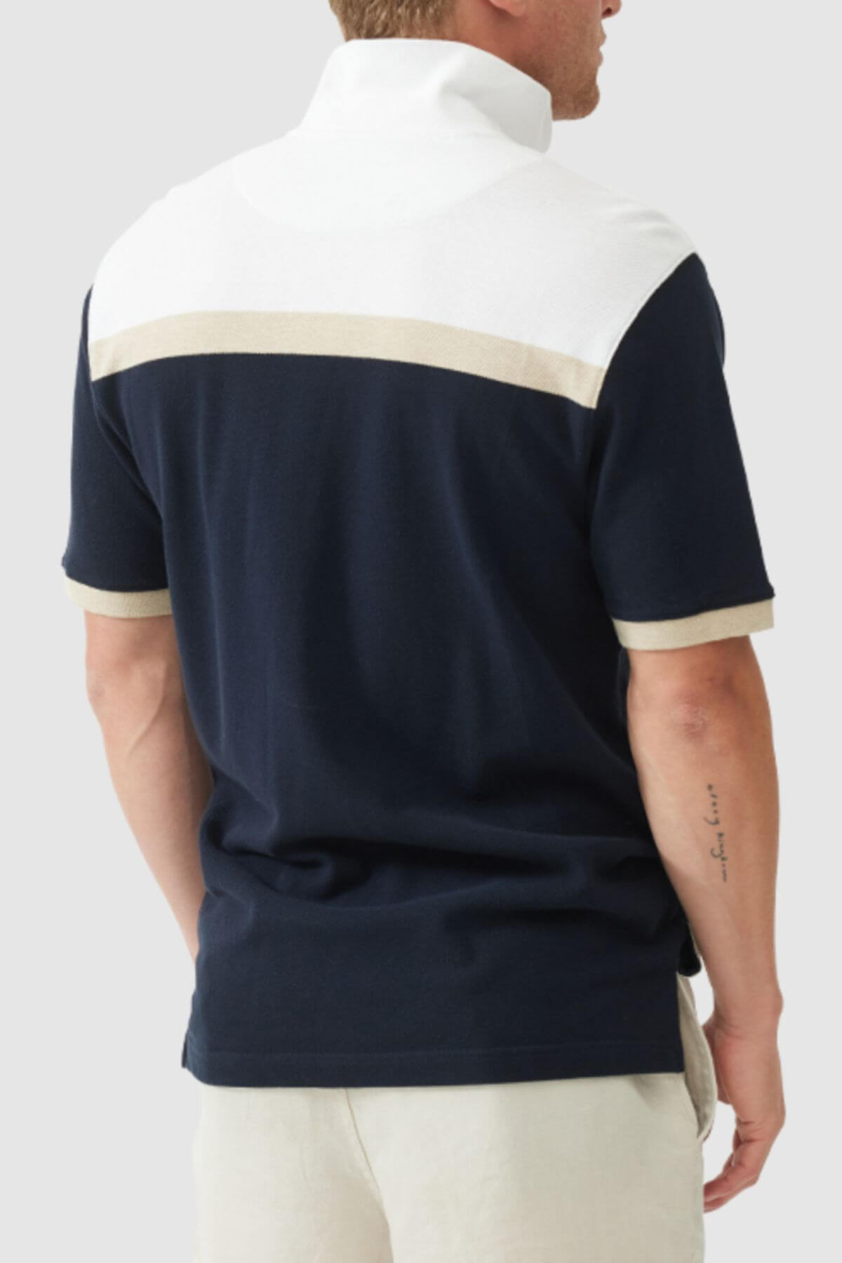 A man is seen from behind in the Rodd & Gunn Whitby Polo, designed with a bold navy body, white upper section, and beige stripes on the back and sleeves. He pairs it with light shorts and displays an arm tattoo.