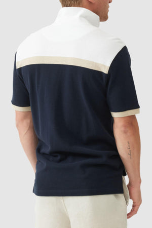 A man is seen from behind in the Rodd & Gunn Whitby Polo, designed with a bold navy body, white upper section, and beige stripes on the back and sleeves. He pairs it with light shorts and displays an arm tattoo.