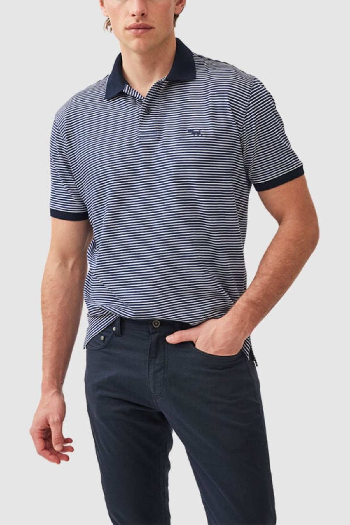 A man wearing the Rodd & Gunn Wye Creek Polo with blue and white stripes, featuring a dark collar and cuffs, paired with dark pants. He has one hand in his pocket and stands against a plain light gray background.