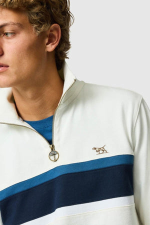 A young person with curly hair wears the Rodd & Gunn Chesterfield Sweat, a cream quarter-zip pullover made from Australian Good Earth Cotton, featuring blue and navy stripes and an embroidered dog logo, layered over a blue shirt.
