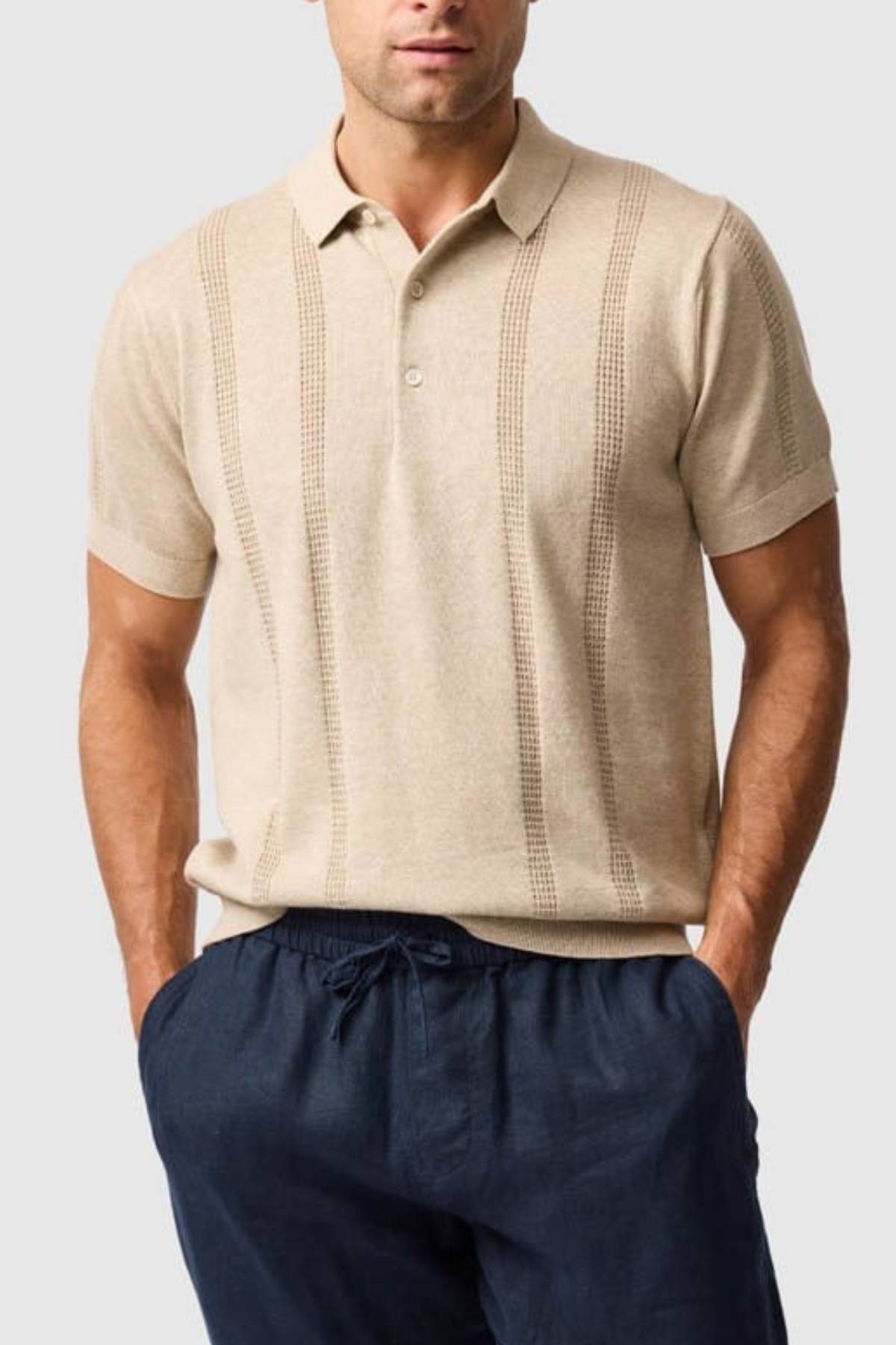 A man wears the Rodd & Gunn Brooklands Knit, a beige Australian cotton polo with vertical textured stripes, and dark blue drawstring pants, standing with hands in pockets against a light gray background.
