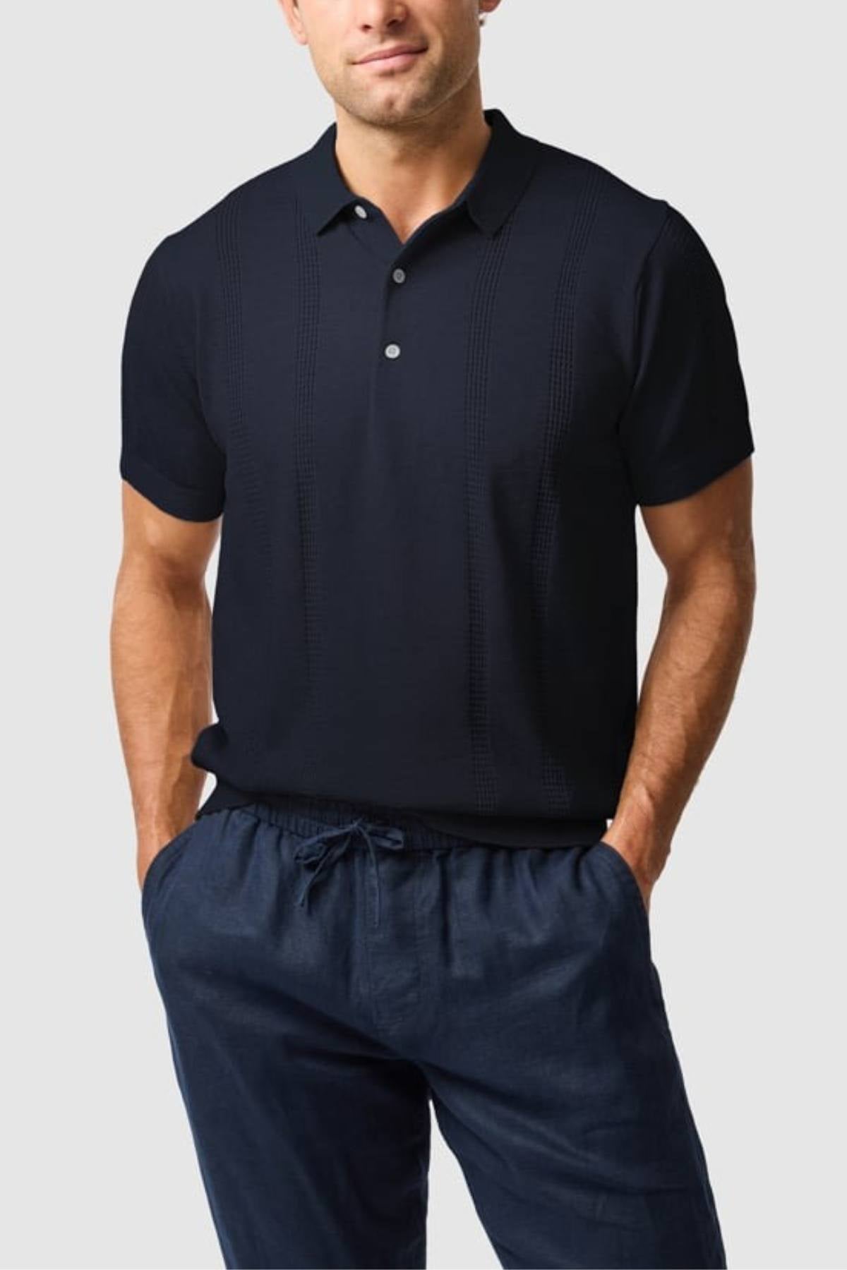 A man stands with his hands in his pockets, wearing the Rodd & Gunn Brooklands Knit—a dark navy short-sleeve polo and matching drawstring pants made from premium Australian cotton—against a light gray background.