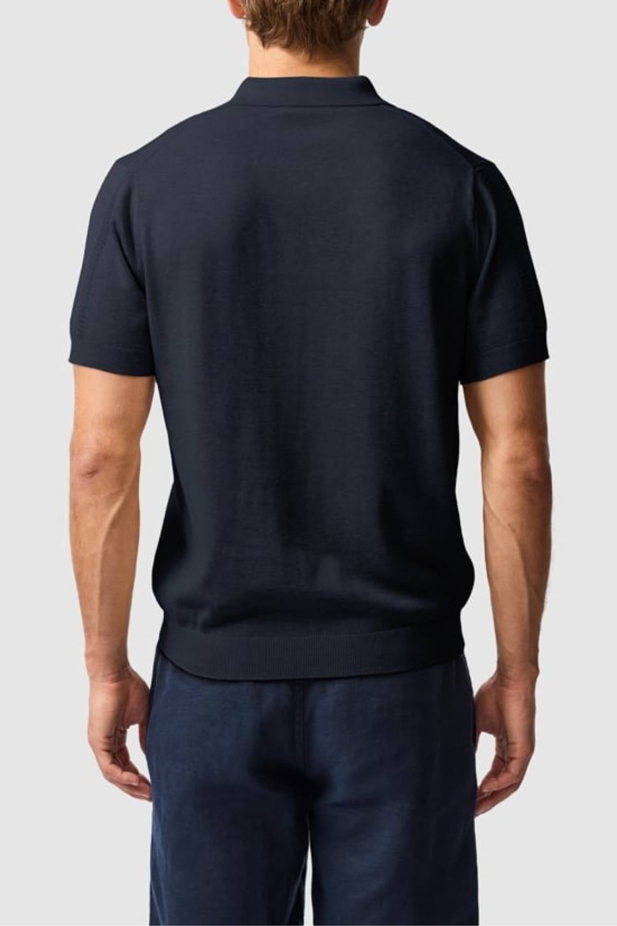 A man is seen from behind wearing the Rodd & Gunn Brooklands Knit short-sleeve dark navy polo in Australian cotton and matching navy pants, standing against a plain light gray background.