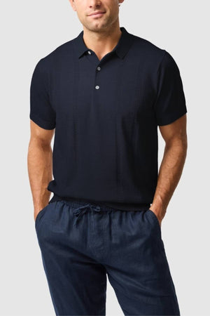 A man stands with his hands in his pockets, wearing the Rodd & Gunn Brooklands Knit—a dark navy short-sleeve polo and matching drawstring pants made from premium Australian cotton—against a light gray background.
