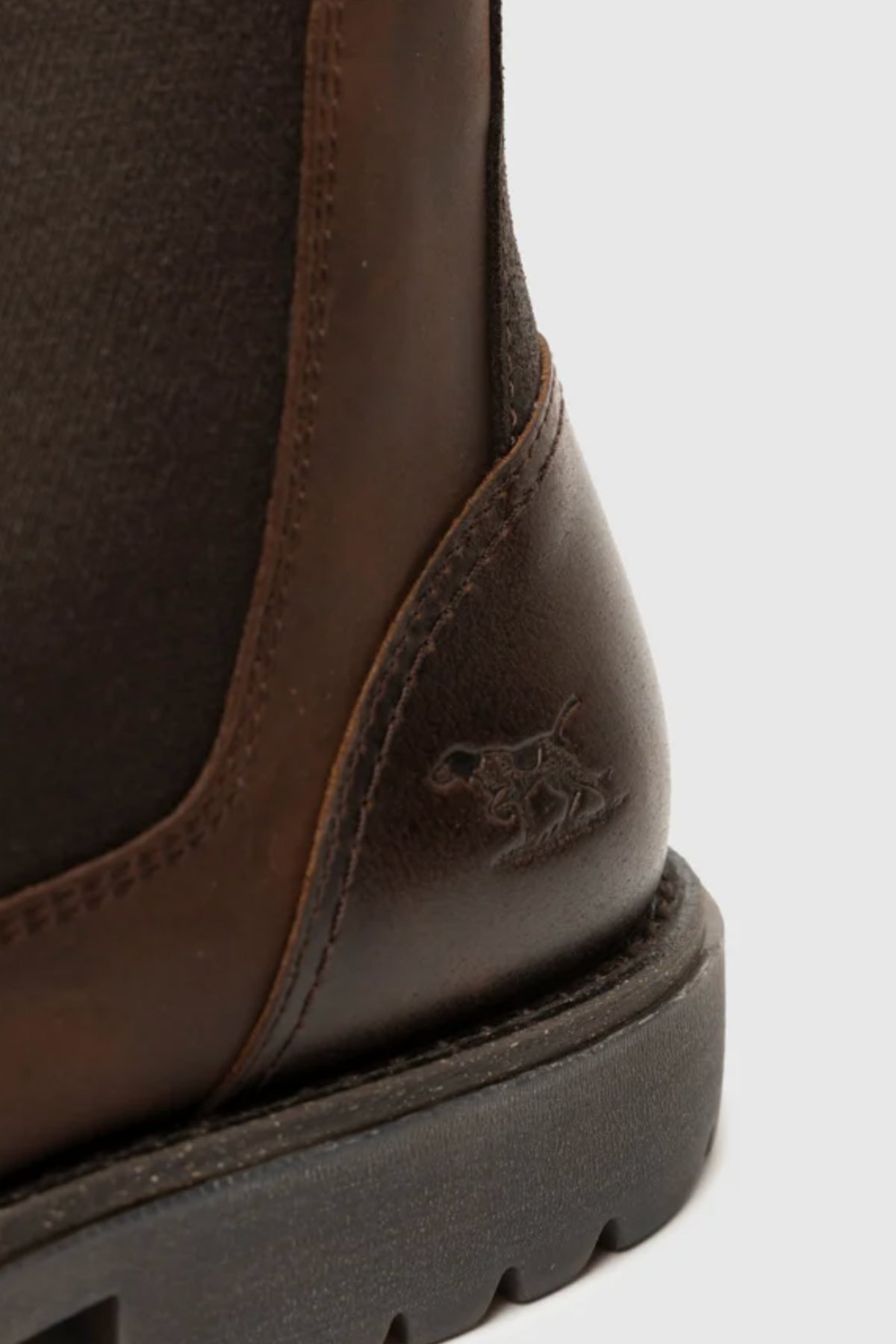 Close-up of the heel on the Rodd & Gunn Clearmont Chelsea Boot in dark brown leather, highlighting visible stitching and a small dog logo embossed near the heel on a gray rubber sole for comfort.