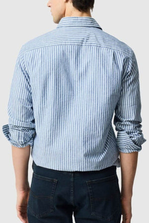 A man is seen from behind wearing the Rodd & Gunn Fernside Sports Fit Shirt, featuring blue and white stripes, rolled-up sleeves, and dark jeans. Made from soft European cotton for a relaxed yet polished appearance.