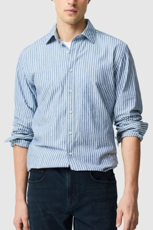A man stands against a light gray background, wearing the Rodd & Gunn Fernside Sports Fit Shirt—light blue and white vertical stripes with rolled-up sleeves—and dark jeans. His face is partially out of frame.