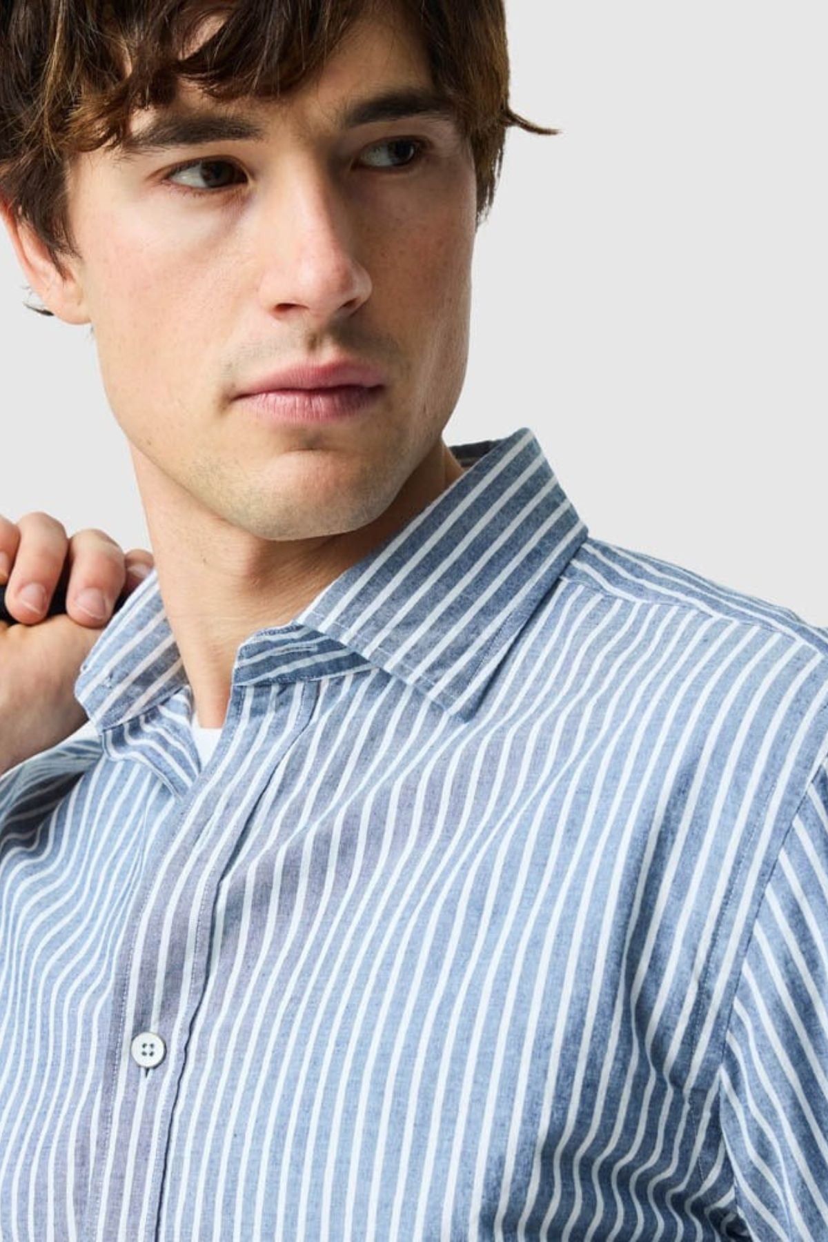 A young man with brown hair looks to the side, holding his shirt collar. He wears a blue and white vertically striped Rodd & Gunn Fernside Sports Fit Shirt, crafted from premium European cotton. The background is light gray.