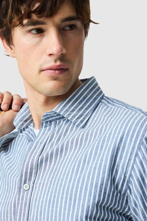 A young man with brown hair looks to the side, holding his shirt collar. He wears a blue and white vertically striped Rodd & Gunn Fernside Sports Fit Shirt, crafted from premium European cotton. The background is light gray.