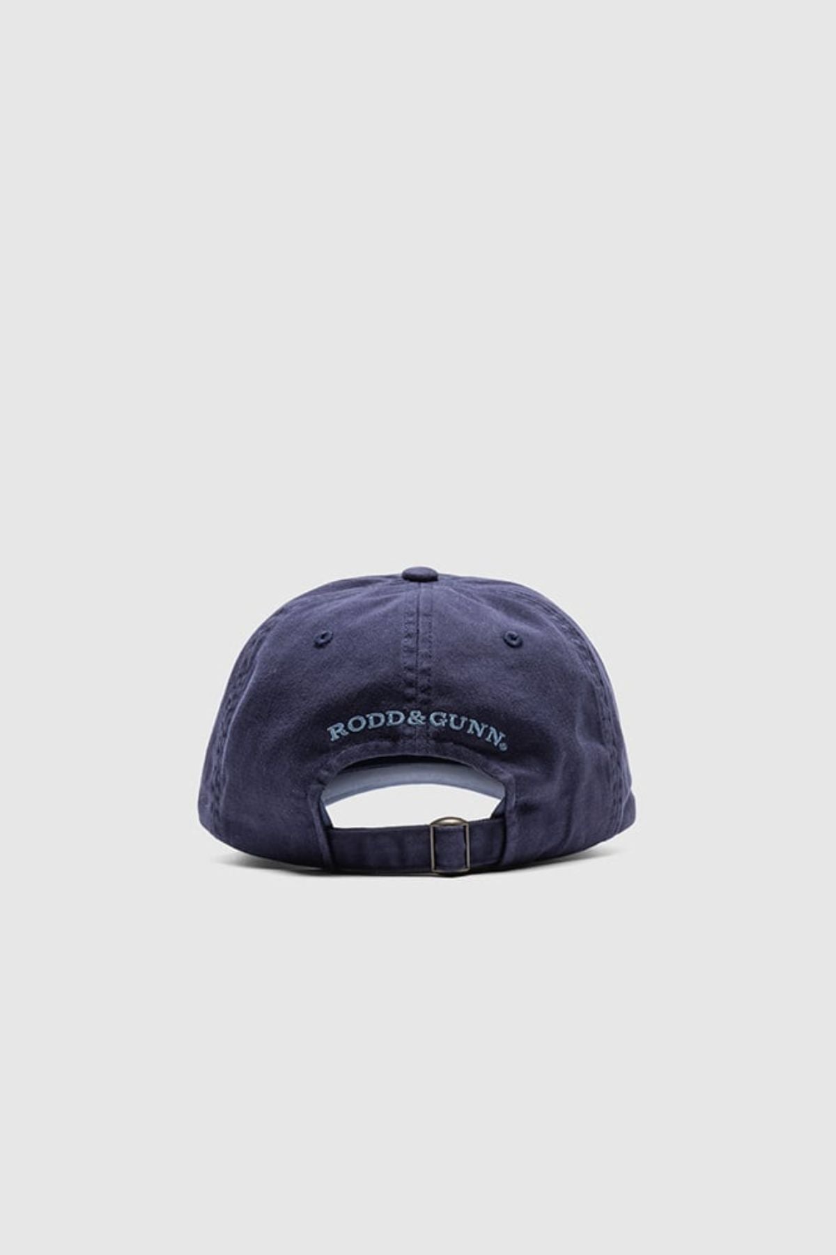 The Rodd & Gunn Gunn Cap, in navy blue cotton twill, is shown from the back with "RODD & GUNN" embroidered above its adjustable strap and buckle. The background is a plain light gray.